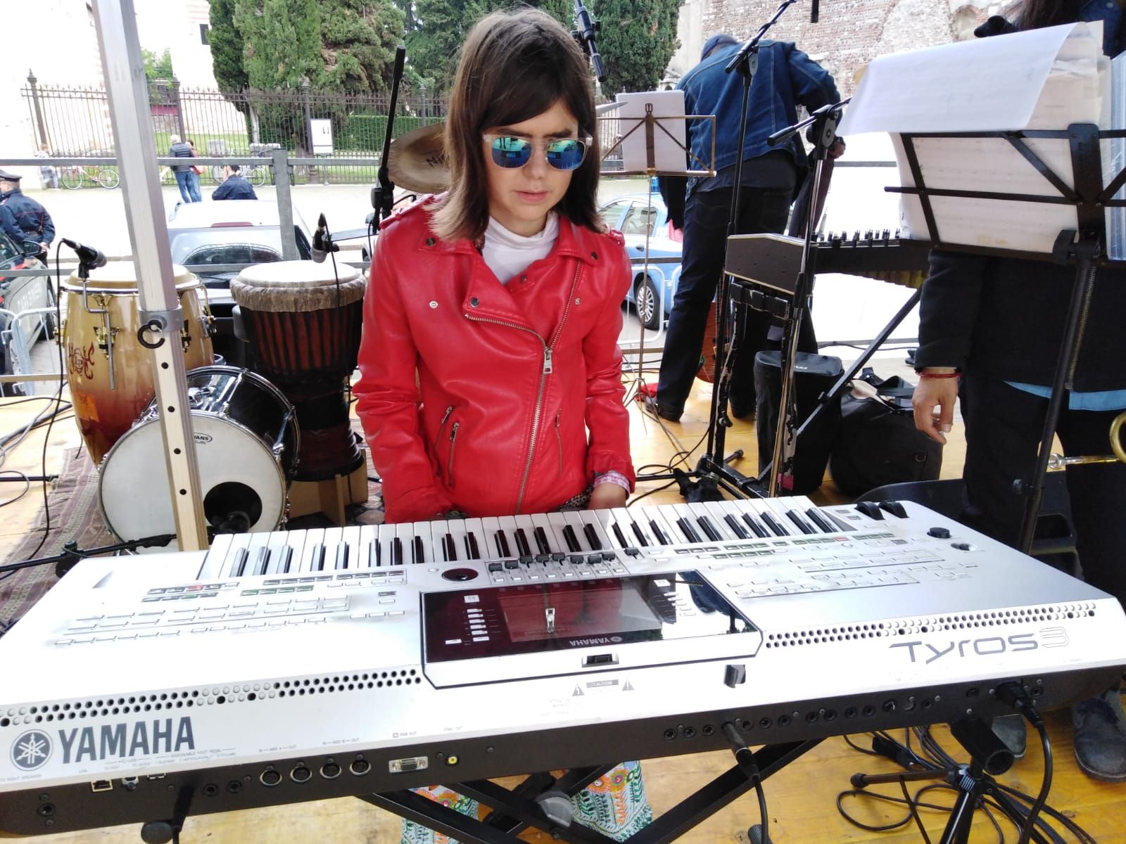 Anna in giacca rossa e occhiali a specchio alla tastiera, su un palco in piazza